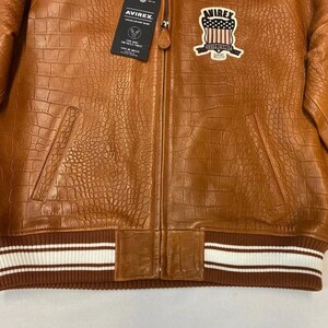 May include: A brown leather jacket with a crocodile texture. The jacket has a zipper closure, two front pockets, and a ribbed hem with white stripes. A patch with a shield design is on the chest. The brand tag is visible.