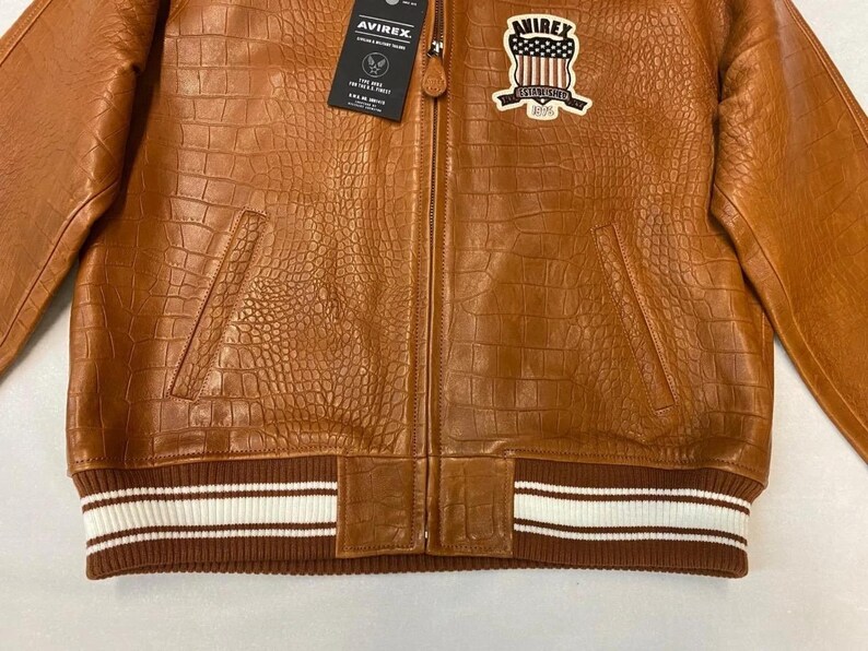 May include: A brown leather jacket with a crocodile texture. The jacket has a zipper closure, two front pockets, and a ribbed hem with white stripes. A patch with a shield design is on the chest. The brand tag is visible.