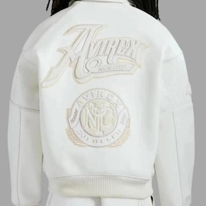 Puede incluir: Chaqueta blanca con el logo "AVIREX NEW YORK" bordado en la espalda. La chaqueta presenta un dobladillo y pu&ntilde;os acanalados, y un parche con las palabras "TRADE AVIREX INC GOLD CLUB". La chaqueta tiene un estilo bomber cl&aacute;sico.