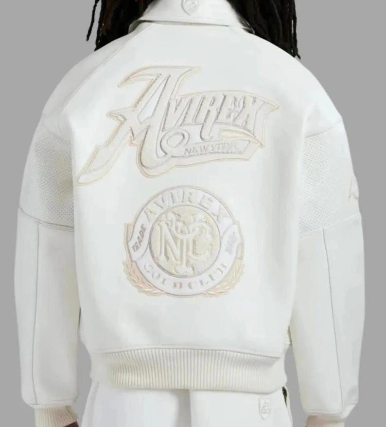 Puede incluir: Chaqueta blanca con el logo "AVIREX NEW YORK" bordado en la espalda. La chaqueta presenta un dobladillo y pu&ntilde;os acanalados, y un parche con las palabras "TRADE AVIREX INC GOLD CLUB". La chaqueta tiene un estilo bomber cl&aacute;sico.