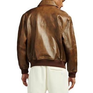 Pu&ograve; includere: Giacca bomber in pelle marrone con colletto e polsini a coste. La giacca ha un design classico con cerniera frontale e una toppa sulla manica superiore sinistra. La giacca &egrave; indossata con pantaloni color crema.