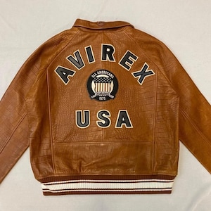 May include: Brown leather jacket with the text "AVIREX" arched across the back, above a patch with "ALL AMERICANS" and "USA". The jacket has a textured pattern and white and brown striped cuffs and hem.