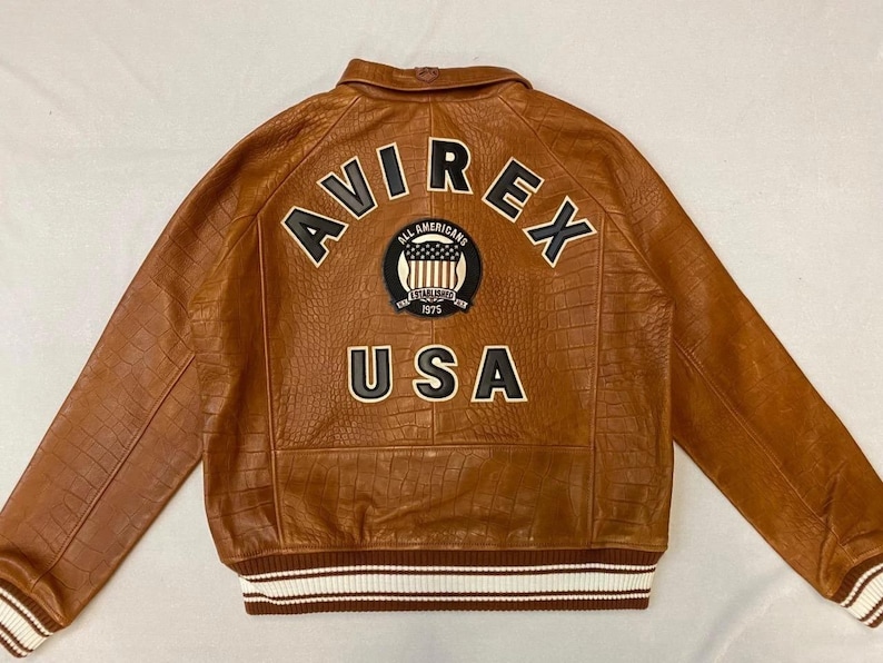 May include: Brown leather jacket with the text "AVIREX" arched across the back, above a patch with "ALL AMERICANS" and "USA". The jacket has a textured pattern and white and brown striped cuffs and hem.