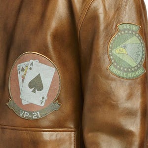 Pu&ograve; includere: Primo piano di una giacca di pelle marrone con due toppe. Una toppa presenta carte da gioco e il testo "VP-21". L'altra toppa ha un disegno di aquila e il testo "Triple Nickel".