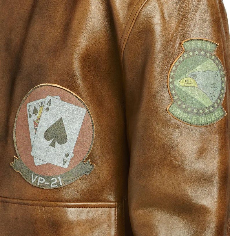 Pu&ograve; includere: Primo piano di una giacca di pelle marrone con due toppe. Una toppa presenta carte da gioco e il testo "VP-21". L'altra toppa ha un disegno di aquila e il testo "Triple Nickel".
