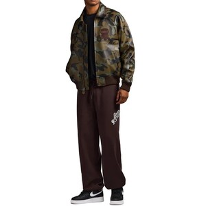 May include: A camouflage-patterned bomber jacket with a brown collar and cuffs. The jacket has a patch on the chest. The person is wearing brown sweatpants with a white logo and black sneakers.