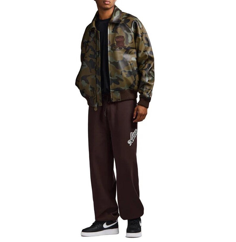 May include: A camouflage-patterned bomber jacket with a brown collar and cuffs. The jacket has a patch on the chest. The person is wearing brown sweatpants with a white logo and black sneakers.