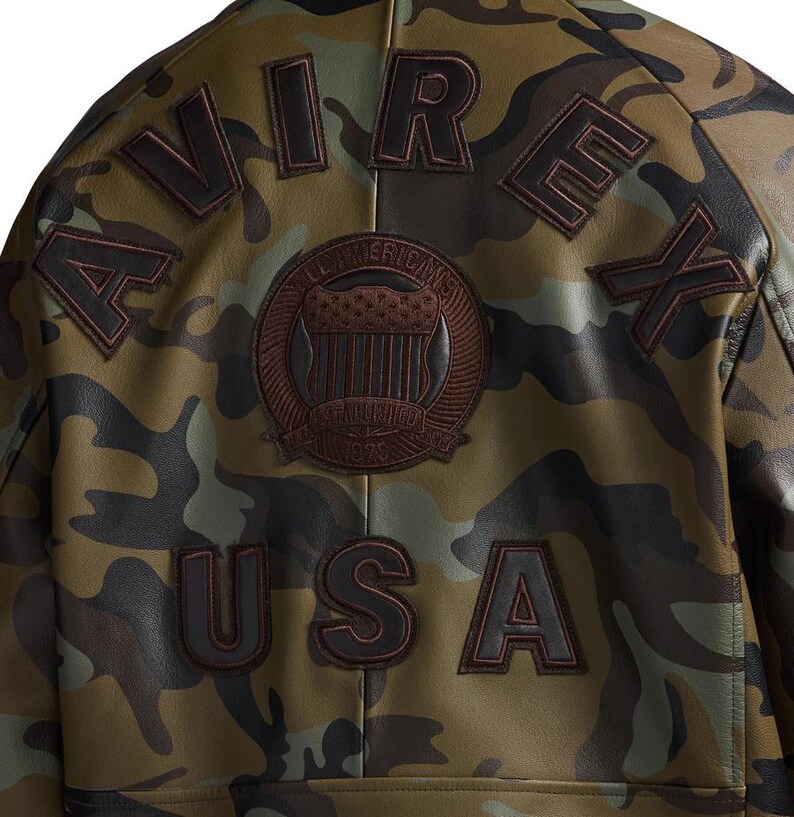 May include: A camouflage-patterned jacket with the word "AVIREX" and "USA" stitched on the back. The jacket features a brown patch with the words "All American's Established 1975". The jacket is in shades of green, brown, and black.