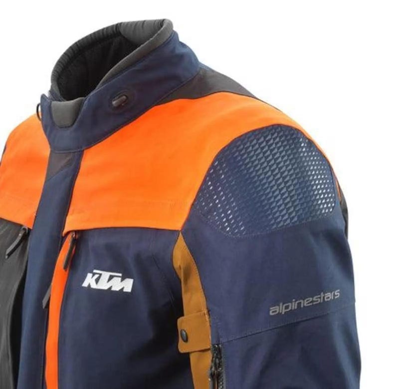 Puede incluir: Chaqueta de moto azul marino y naranja con el logotipo de KTM y la marca Alpinestars. La chaqueta presenta un cuello alto, un panel de malla en el hombro y un bolsillo con cremallera. La chaqueta est&aacute; dise&ntilde;ada para montar en moto.