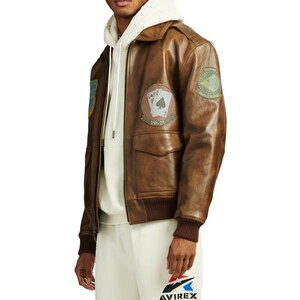 Pu&ograve; includere: Giacca bomber in pelle marrone con toppe, tra cui un disegno di carte da gioco e la scritta "VP-21". La giacca ha colletto, polsini e orlo a coste. La giacca &egrave; indossata sopra una felpa con cappuccio bianca.
