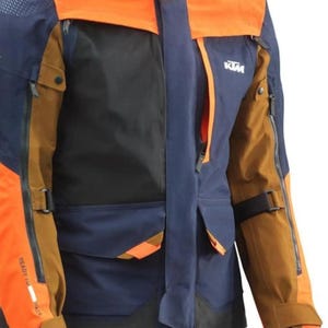 Puede incluir: Primer plano de una chaqueta de moto en tonos naranja, azul marino, negro y marr&oacute;n. La chaqueta tiene m&uacute;ltiples bolsillos, cremalleras y el logotipo de KTM. Las mangas tienen correas ajustables y el texto "READY TO RACE".