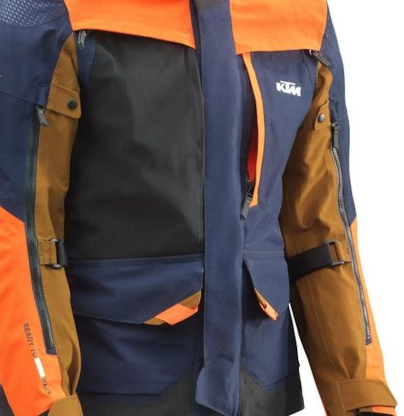 Puede incluir: Primer plano de una chaqueta de moto en tonos naranja, azul marino, negro y marr&oacute;n. La chaqueta tiene m&uacute;ltiples bolsillos, cremalleras y el logotipo de KTM. Las mangas tienen correas ajustables y el texto "READY TO RACE".