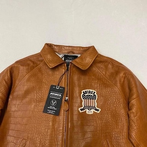May include: A brown leather jacket with a crocodile texture. The jacket has a zipper closure, a collar, and a patch on the chest with the text "AVIREX ESTABLISHED 1975". A tag with the "AVIREX" logo hangs from the zipper.