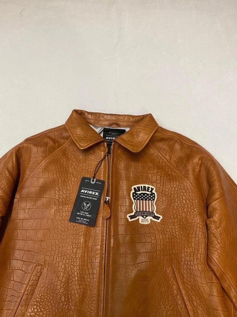 May include: A brown leather jacket with a crocodile texture. The jacket has a zipper closure, a collar, and a patch on the chest with the text "AVIREX ESTABLISHED 1975". A tag with the "AVIREX" logo hangs from the zipper.