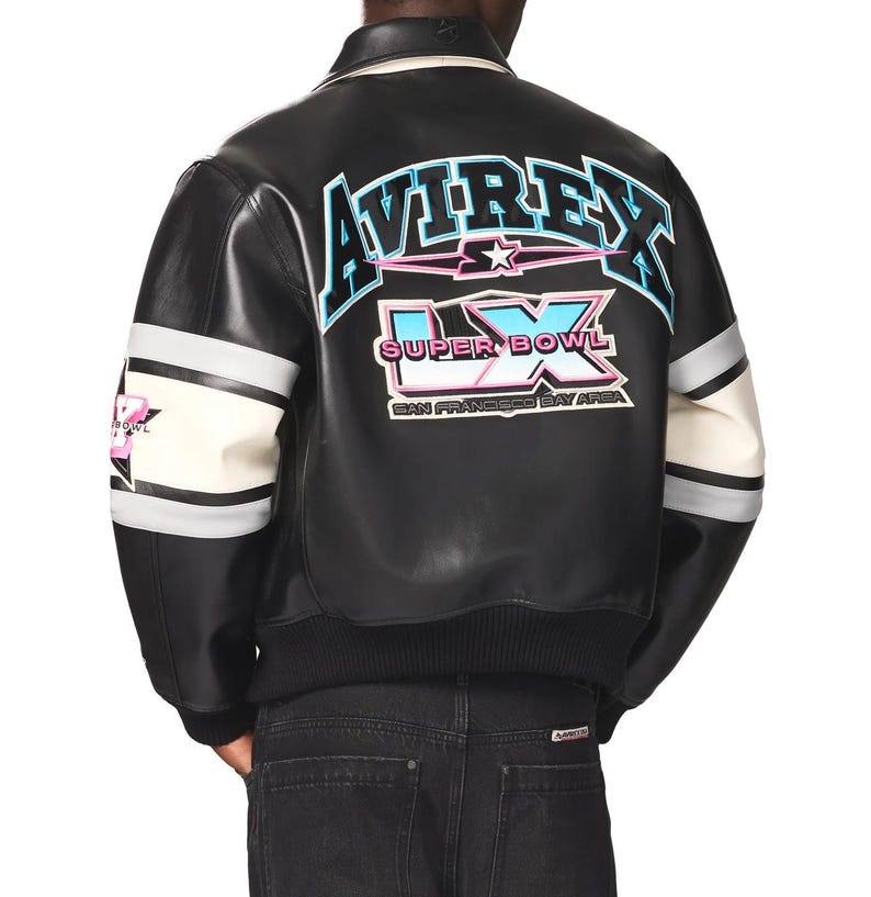 May include: Black leather bomber jacket with white and gray striped sleeves. The back features the text "AVIREX LX SUPER BOWL SAN FRANCISCO BAY AREA" in blue, pink, and white. The jacket has a black ribbed hem and collar.