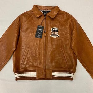 May include: A brown leather jacket with a crocodile texture. The jacket has a zip closure, a collar, and a patch on the chest. The cuffs and hem are ribbed with white stripes. The brand tag reads "AVIREX".