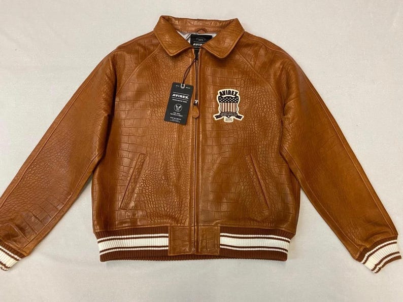 May include: A brown leather jacket with a crocodile texture. The jacket has a zip closure, a collar, and a patch on the chest. The cuffs and hem are ribbed with white stripes. The brand tag reads "AVIREX".