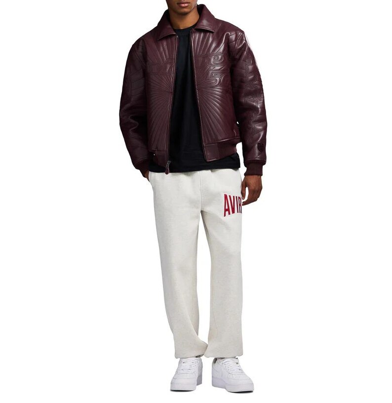 May include: A burgundy leather jacket with a quilted design and a black t-shirt. The person is wearing light gray sweatpants with the word "AVIR" in red letters. White sneakers complete the outfit.