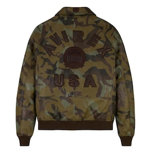 May include: Camouflage-patterned leather jacket with the word "AVIREX" arched across the shoulders and "USA" below. The jacket features a brown ribbed hem and cuffs, and a collar. The jacket is a military-inspired style.
