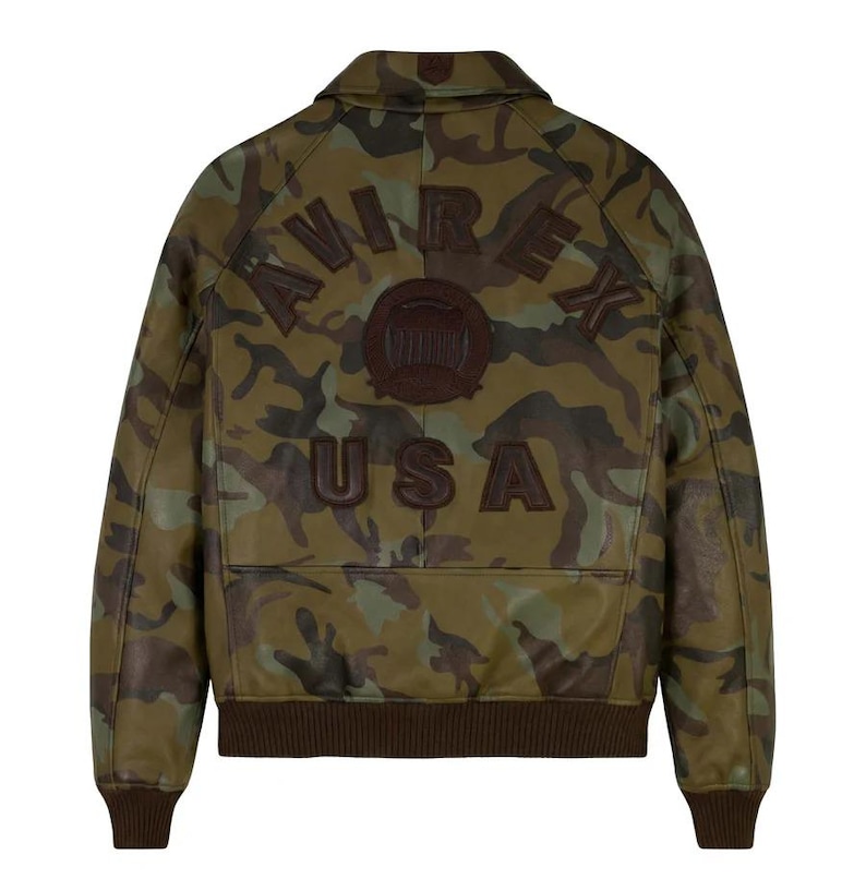 May include: Camouflage-patterned leather jacket with the word "AVIREX" arched across the shoulders and "USA" below. The jacket features a brown ribbed hem and cuffs, and a collar. The jacket is a military-inspired style.