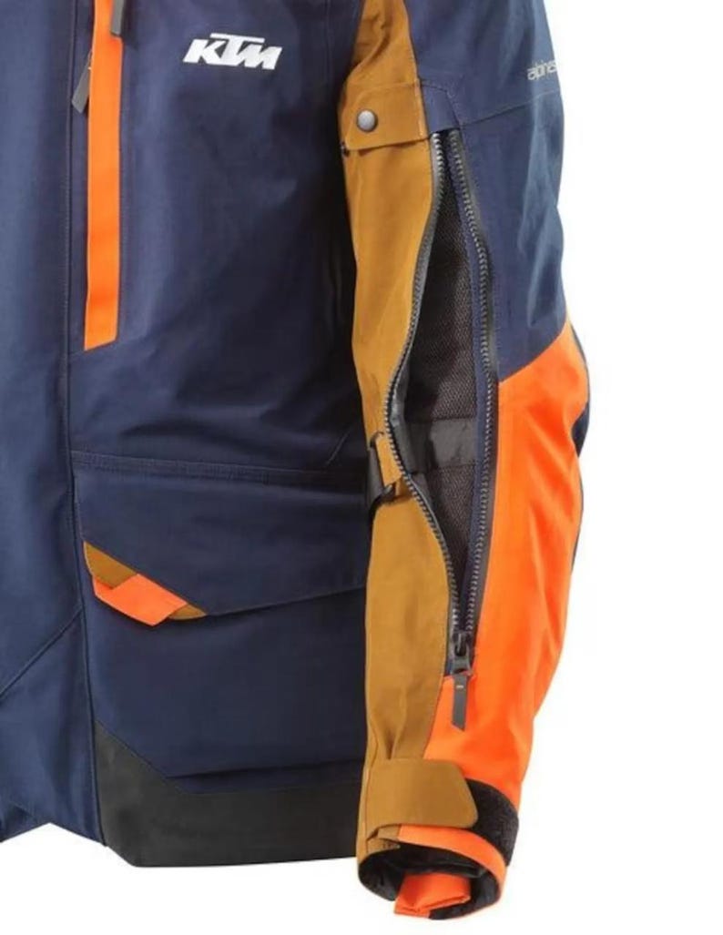 Puede incluir: Chaqueta de moto azul marino y naranja con el logo KTM. La chaqueta presenta detalles en naranja en las mangas y los bolsillos, con una abertura con cremallera en la manga. La chaqueta tiene cuello y pu&ntilde;os negros.