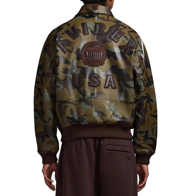 May include: Camouflage-patterned leather jacket with the text "AVIREX USA" on the back. The jacket features a brown ribbed collar, cuffs, and hem. The camouflage design incorporates shades of green, brown, and black.