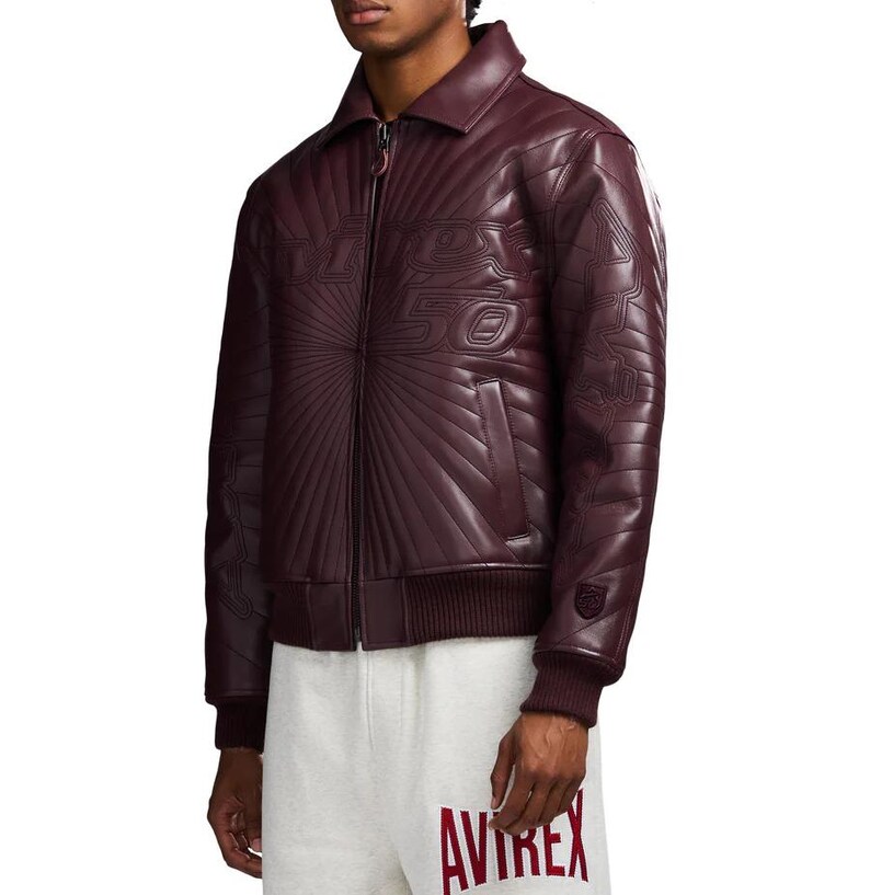 May include: A burgundy leather jacket with a zip-up front, collar, and ribbed cuffs. The jacket features embossed text and a sunburst design on the front. The sleeves have a quilted pattern. The jacket is worn with gray sweatpants.