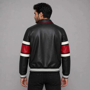 Pu&ograve; includere: Giacca in pelle nera con maniche a righe rosse e bianche e orlo a righe bianche e nere. La giacca ha un colletto rosso e un classico design da bomber. Il modello indossa jeans neri.
