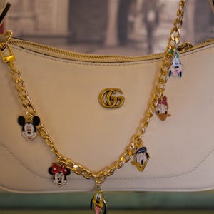 May include: Cream-colored handbag with a gold chain strap adorned with cartoon character charms. The charms include Mickey Mouse, Minnie Mouse, Donald Duck, and Pluto. The bag features a gold double-G logo.