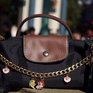 Peut inclure: Sac &agrave; main noir avec rabat et poign&eacute;e en cuir marron. Une cha&icirc;ne dor&eacute;e avec des breloques coquillages et personnages de dessins anim&eacute;s pend de la sacoche. Le sac a une fermeture &agrave; bouton.