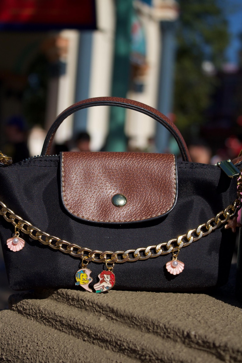 Peut inclure: Sac &agrave; main noir avec rabat et poign&eacute;e en cuir marron. Une cha&icirc;ne dor&eacute;e avec des breloques coquillages et personnages de dessins anim&eacute;s pend de la sacoche. Le sac a une fermeture &agrave; bouton.