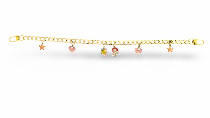 Peut inclure: Bracelet &agrave; cha&icirc;ne dor&eacute;e avec plusieurs breloques. Les breloques comprennent une &eacute;toile de mer, un coquillage et un charm sur le th&egrave;me de la sir&egrave;ne. Le bracelet est pr&eacute;sent&eacute; sur un fond blanc.