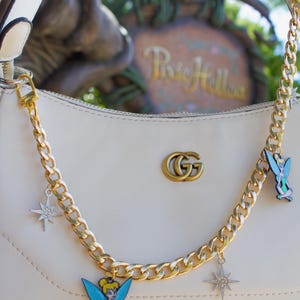 May include: Cream-colored handbag with a gold chain strap and decorative charms. The charms include a starburst and a cartoon fairy. The bag features a gold double-G logo and gold hardware.