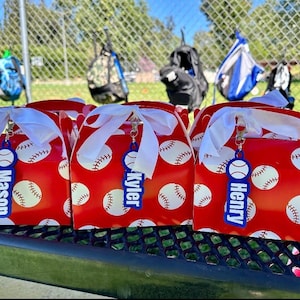 Pu&ograve; includere: Tre scatole rosse con motivo a palline da baseball bianche, ciascuna legata con un nastro bianco e dotata di un'etichetta in acrilico blu con i nomi Mason, Kyler e Henry stampati in bianco, fissata con un moschettone in metallo argentato.