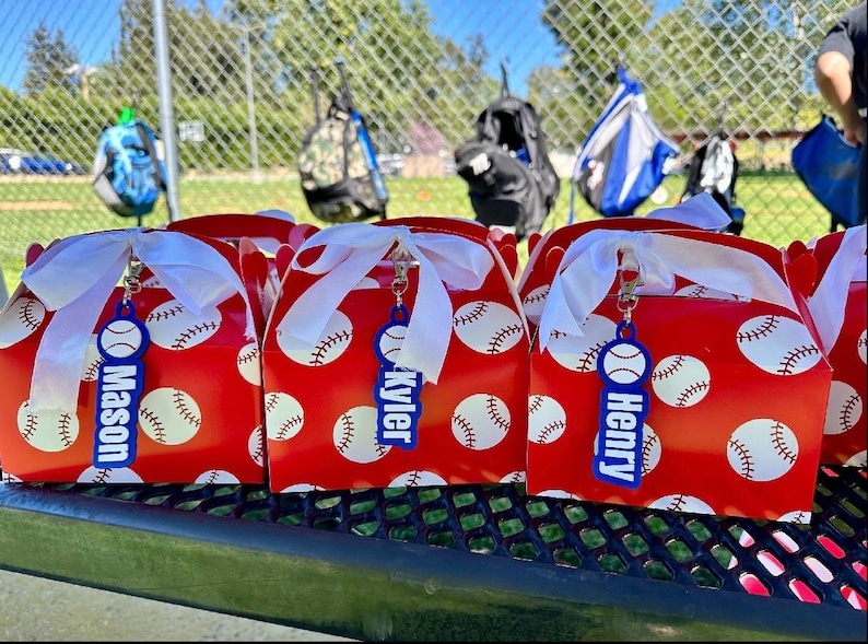 Pu&ograve; includere: Tre scatole rosse con motivo a palline da baseball bianche, ciascuna legata con un nastro bianco e dotata di un'etichetta in acrilico blu con i nomi Mason, Kyler e Henry stampati in bianco, fissata con un moschettone in metallo argentato.