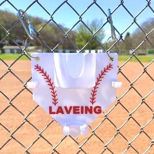 May include: A white baseball-shaped item with red stitching and the word "LAVEING" in red, hanging from a chain-link fence. The item has a curved top and two small white holders. The background shows a baseball field.