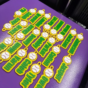 Pu&ograve; includere: Una collezione di piccole etichette in plastica gialla personalizzate, ognuna con una grafica di baseball bianca con cuciture rosse e un nome verde stampato in un carattere sans-serif in grassetto. Ogni etichetta include un piccolo occhiello.