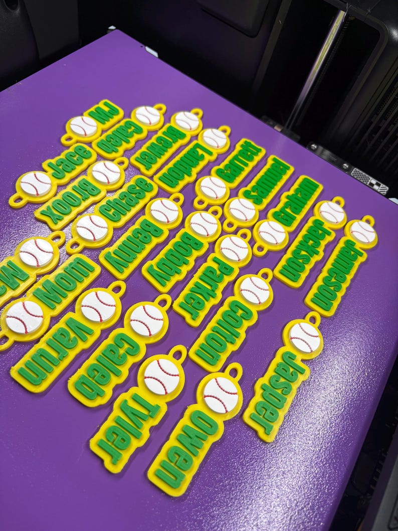 Pu&ograve; includere: Una collezione di piccole etichette in plastica gialla personalizzate, ognuna con una grafica di baseball bianca con cuciture rosse e un nome verde stampato in un carattere sans-serif in grassetto. Ogni etichetta include un piccolo occhiello.