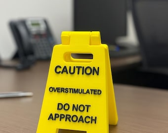 May include: A bright yellow caution sign with black text that reads "CAUTION OVERSTIMULATED DO NOT APPROACH." The sign is in a triangular shape and is placed on a wooden desk in an office setting.