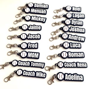 Pu&ograve; includere: Una collezione di portachiavi personalizzati a tema baseball, ognuno caratterizzato da un'icona di palla da baseball bianca con cuciture rosse attaccata a un'etichetta rettangolare scura. Le etichette mostrano vari nomi tra cui Damian, Menuah, Mickey, Jaime, Jacob, Fred, MOM, Coach Tommy, Coach Mike, Noel, Logan, Dylan, Joshua, Andrew, Luca, Roman, Coach Rena e Adelina in lettere bianche.