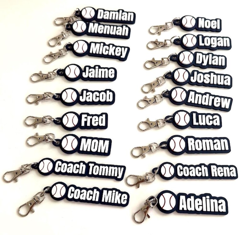 Pu&ograve; includere: Una collezione di portachiavi personalizzati a tema baseball, ognuno caratterizzato da un'icona di palla da baseball bianca con cuciture rosse attaccata a un'etichetta rettangolare scura. Le etichette mostrano vari nomi tra cui Damian, Menuah, Mickey, Jaime, Jacob, Fred, MOM, Coach Tommy, Coach Mike, Noel, Logan, Dylan, Joshua, Andrew, Luca, Roman, Coach Rena e Adelina in lettere bianche.