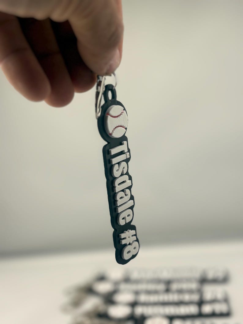 Pu&ograve; includere: Un portachiavi in gomma di colore scuro con un'icona di una palla da baseball bianca nella parte superiore e il testo "Tisdale #8" in lettere bianche in rilievo. Il portachiavi &egrave; fissato a un anello in metallo argentato per le chiavi.