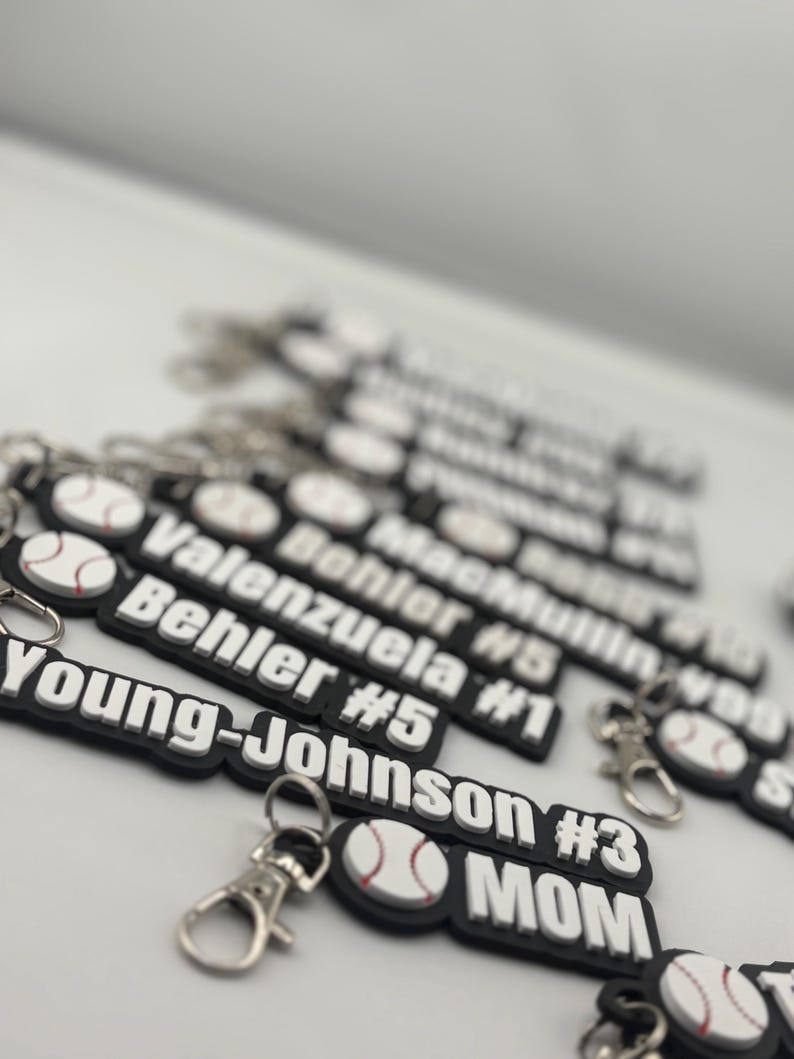 Pu&ograve; includere: Una collezione di portachiavi rettangolari bianchi e neri con una grafica da baseball e testo personalizzato che include nomi come Young-Johnson #3, Behler #5, Valenzuela #1 e MacMullin #99, ciascuno fissato a un moschettone in metallo argentato.