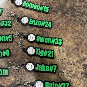 Pu&ograve; includere: Una collezione di portachiavi in gomma nera e verde neon, ognuno con una grafica di baseball bianca e un nome personalizzato con numero di maglia, inclusi Roman#15, Enzo#24, Owen#33, Tig#21, Jake#7 e Nate#27, fissati a moschettoni in metallo argentato.