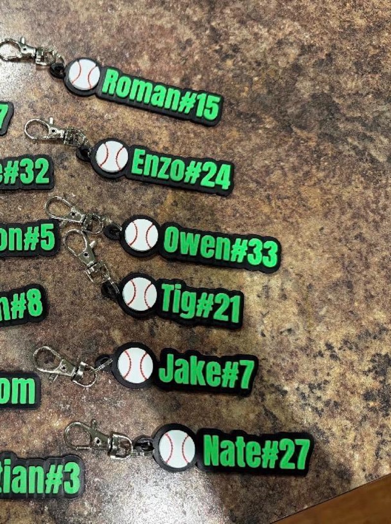 Pu&ograve; includere: Una collezione di portachiavi in gomma nera e verde neon, ognuno con una grafica di baseball bianca e un nome personalizzato con numero di maglia, inclusi Roman#15, Enzo#24, Owen#33, Tig#21, Jake#7 e Nate#27, fissati a moschettoni in metallo argentato.