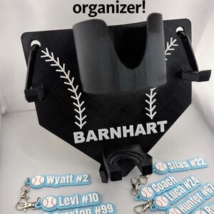 Pu&ograve; includere: Un organizer nero per baseball e softball a forma di casa base con portabicchieri centrale, due ganci laterali e il nome BARNHART stampato in bianco. Sotto ci sono diversi portachiavi rettangolari azzurri con grafiche di palline da baseball.