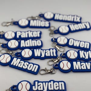 May include: A collection of blue and white baseball-themed keychains. Each keychain features a baseball design and a name in white lettering. The keychains have silver-colored clasps.