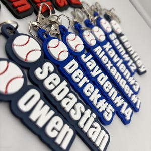 Pu&ograve; includere: Una collezione di portachiavi in silicone blu navy e bianco con un'icona da baseball in rilievo, nomi personalizzati e numeri di maglia. Ogni portachiavi include un moschettone in metallo argentato fissato a un anello circolare.