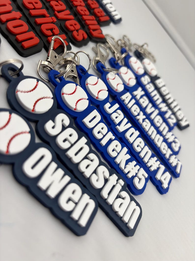 Pu&ograve; includere: Una collezione di portachiavi in silicone blu navy e bianco con un'icona da baseball in rilievo, nomi personalizzati e numeri di maglia. Ogni portachiavi include un moschettone in metallo argentato fissato a un anello circolare.