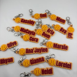Pu&ograve; includere: Una collezione di portachiavi personalizzati gialli e marroni, ognuno con un'icona di palla da softball e un nome o titolo tra cui Kassidy, Emi, Mom, MaCayla, Aria, Dom, Ava, Ari, Olive, Cora, Ana'Jaylee, Marsie, Rhyan, Gianna, Lorelei e Heidi.
